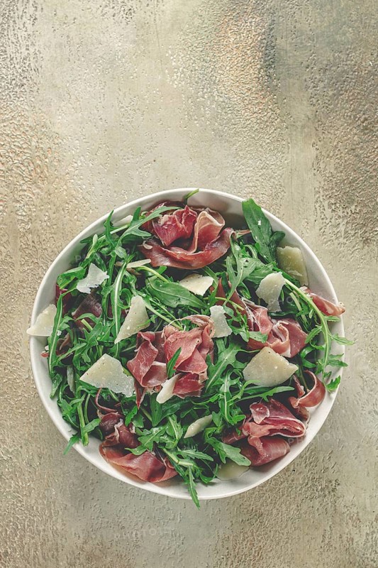 Fresh arugula salad with prosciutto and grated parmesan cheese, on a plate, homemade, no people