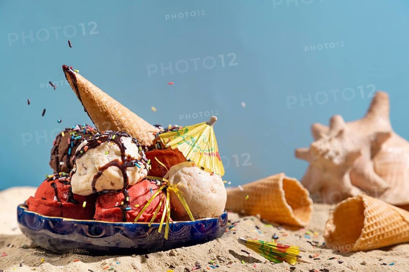 Various flavours of ice cream with chocolate sauce, crumble and parasol on sand with shells
