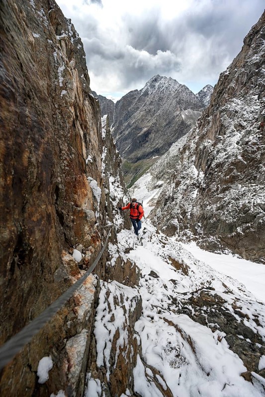 Mountaineer on a rope-secured path in a steep rocky mountain landscape with fresh snow in summer, behind mountain peaks Großer Friedrichskopf and Georgskopf, ascent to Hornscharte, Schobergruppe, Hohe Tauern National Park, Carinthia, Austria