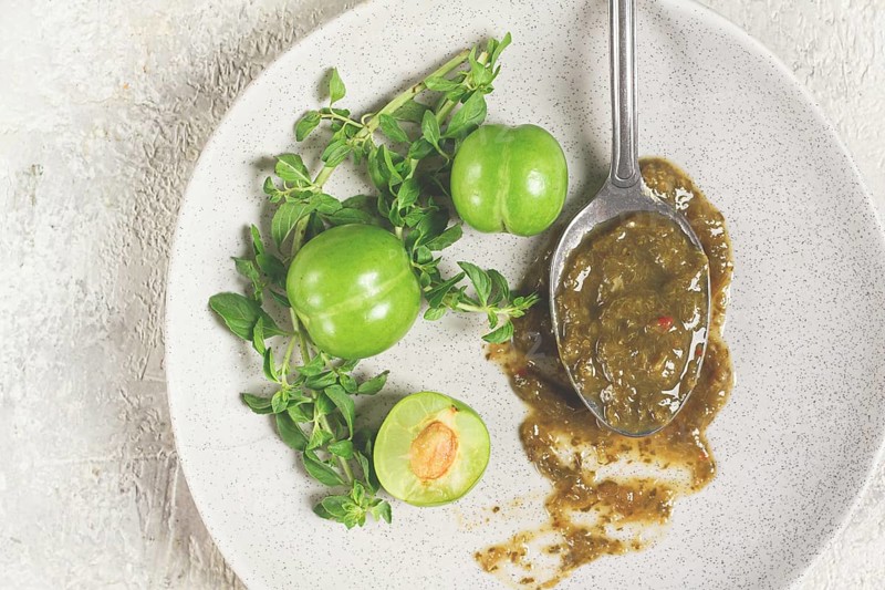 A spoon with tkemali sauce, on a plate, with cherry plum and mint, top view, no people