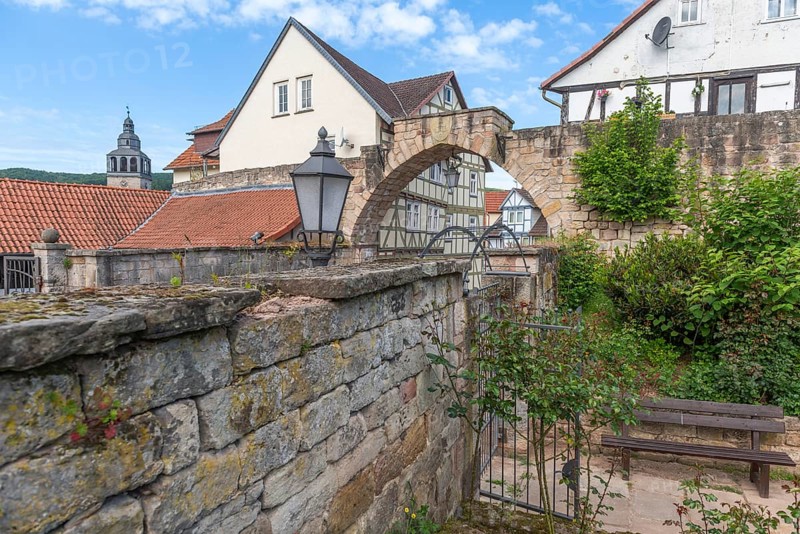 Former stone gate, town gate of the medieval town wall, Bad Sooden-Allendorf, district Allendorf, half-timbered houses, tower of St. Crucis church, bench, lamp, bushes, blue sky, clouds, Werra-Meissner district, Hesse, Germany