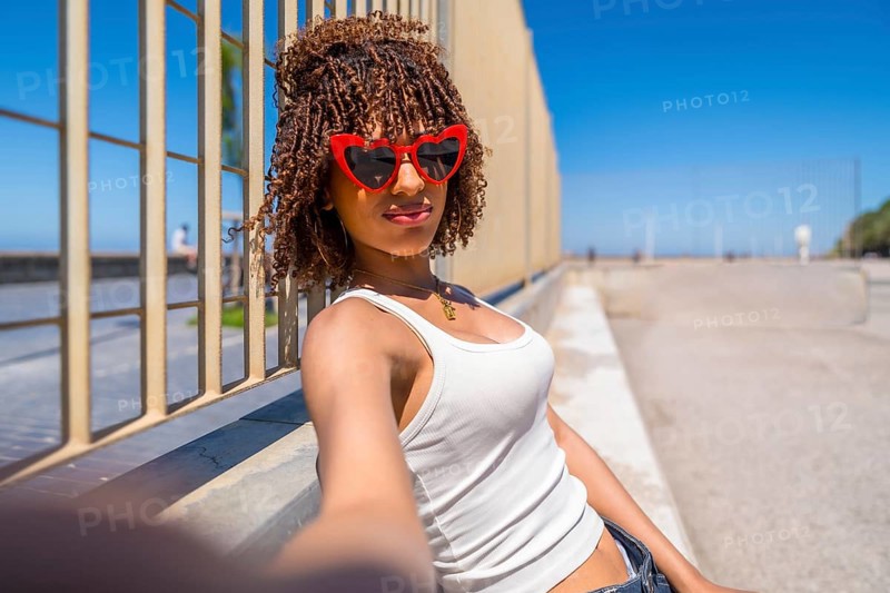 Fashionable young woman with curly hair enjoying a sunny day, taking a selfie by a stylish metal fence, radiating confidence and happiness