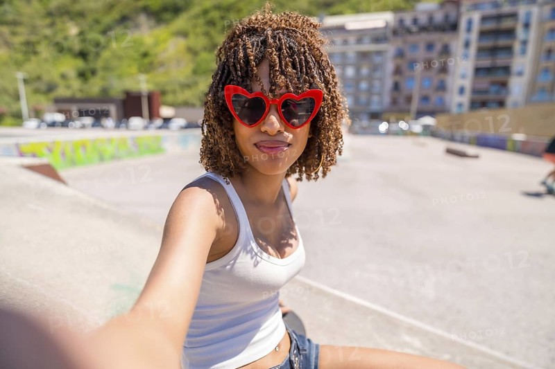 Young woman with heart shaped sunglasses enjoying a sunny day at a skatepark, capturing the moment with a cheerful selfie