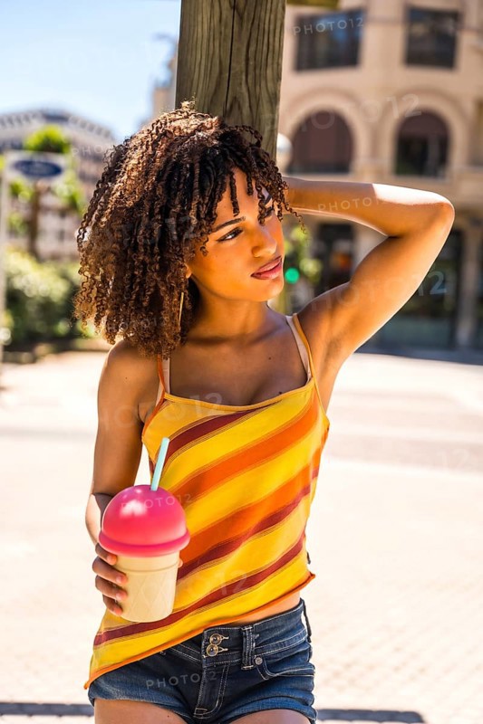 Stylish young woman with curly hair enjoys a cold beverage on a sunny summer day, exuding a carefree and vibrant spirit
