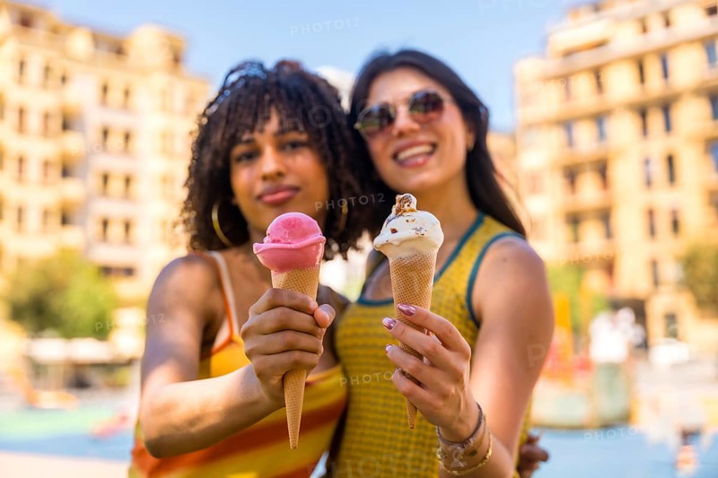 Two young women smiling brightly while holding colorful ice cream cones on a sunny summer day in the vibrant city, enjoying their time together
