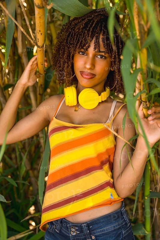 Young woman with yellow headphones enjoying music in a bamboo forest, wearing a colorful striped top and jeans