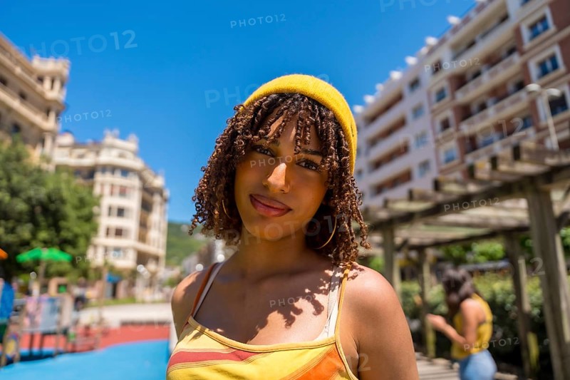 Portrait of a fashionable young woman with curly hair and yellow beanie enjoying a sunny day in an urban park