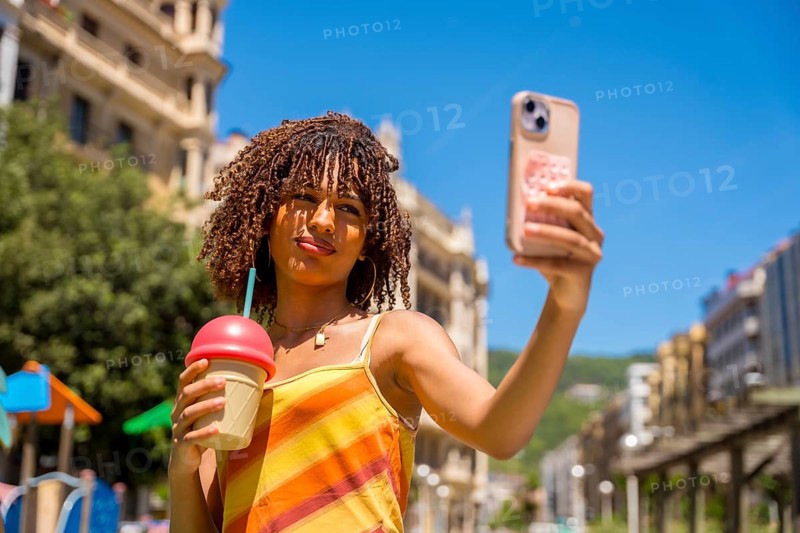 Stylish young woman captures a selfie while holding a refreshing beverage, enjoying a sunny day in a vibrant urban setting