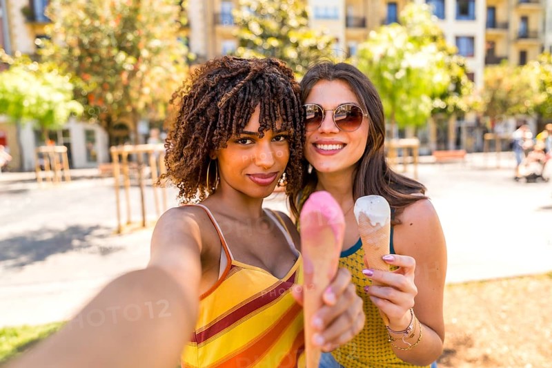 Two cheerful young women enjoying a sunny day in the city park, taking a selfie and showing their delicious ice cream cones