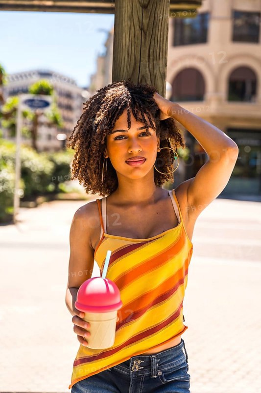 Fashionable young woman enjoying a sunny day in the city, holding a refreshing beverage and radiating happiness while relaxing outdoors