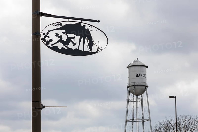 Bandera water tower behind artistic metal sign showing cowboy life on pole in Bandera, Texas, USA