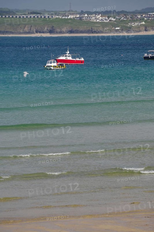 A red boat floats on calm blue water near a wide sandy beach, St Ives, Cornwall, England, United Kingdom