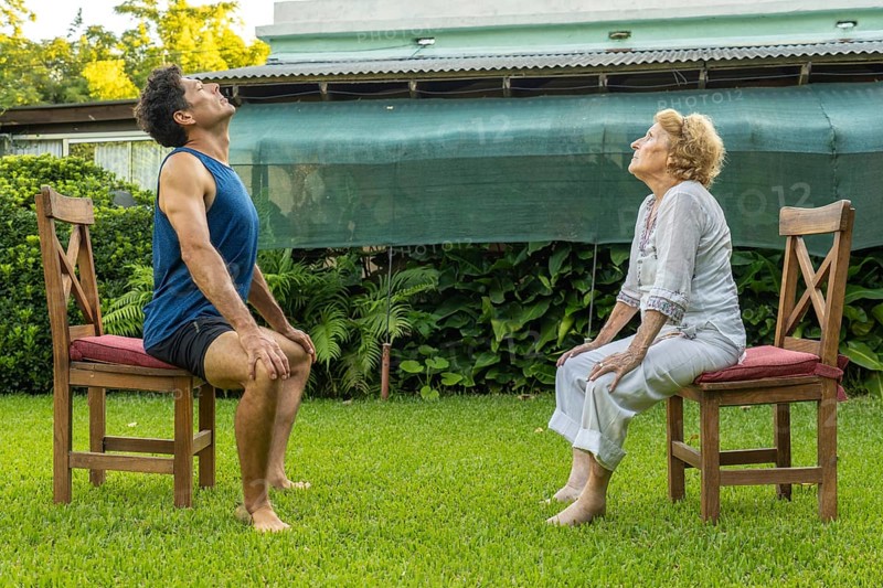 Senior woman and personal trainer engaging in seated exercises on chairs in a serene garden, supporting healthy aging and enhancing mobility through outdoor fitness activities