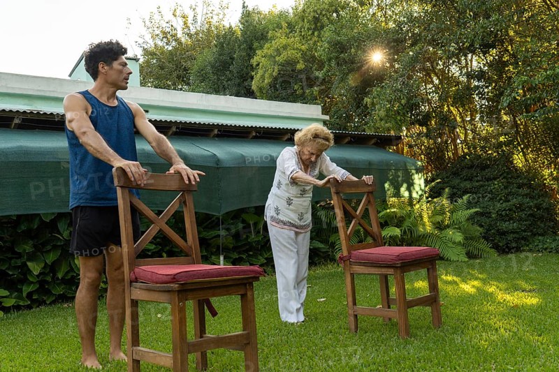 Senior woman practicing physical therapy exercises with the help of her personal trainer, using chairs as support in a backyard setting, promoting health and mobility in old age