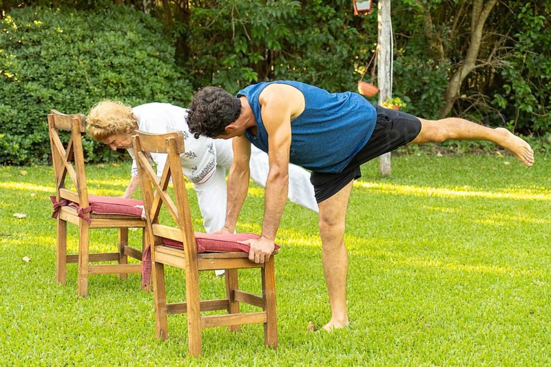 Senior man and woman practicing yoga in garden, doing balancing pose using chairs for support, promoting healthy lifestyle and fitness in old age