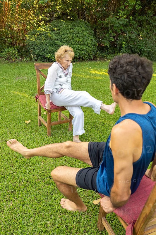 Elderly woman exercising her legs with the help of a physiotherapist, sitting on chairs in a backyard, promoting health and mobility in old age
