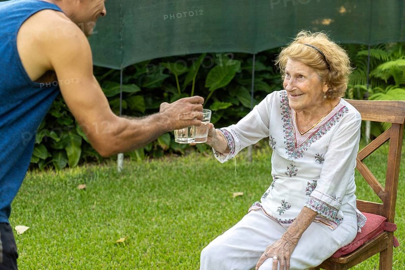 Young male nurse is offering a glass of fresh water to a smiling elderly woman sitting on a wooden chair in a garden, showing care and support for the elderly