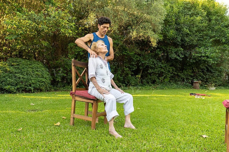 Physiotherapist is giving a shoulder massage to a senior woman sitting on a chair in a peaceful garden setting, promoting relaxation and well-being