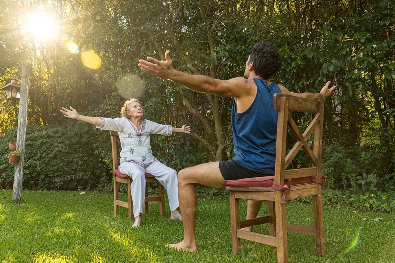 Senior woman practicing seated exercises with the assistance of a physiotherapist in a green backyard, promoting health and mobility in old age