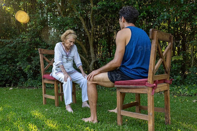Senior woman experiencing knee pain receiving assistance from a physiotherapist during an outdoor therapy session in a peaceful garden setting, promoting health and well-being in old age