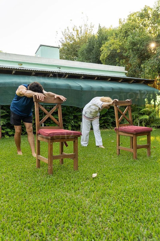 Senior woman and her physiotherapist are bending forward, holding onto chairs in a garden, performing stretching exercises to improve flexibility and mobility