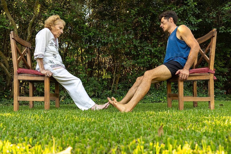 Elderly woman and her personal trainer practicing leg strengthening exercises using chairs in a garden, promoting healthy aging and mobility
