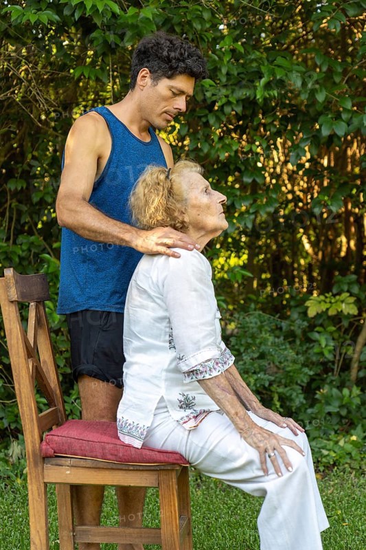 Physiotherapist is guiding a senior woman through posture exercises, providing support and gentle adjustments as she sits on a chair in a serene outdoor setting, promoting mobility and well-being