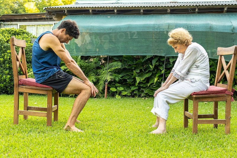 Senior woman and her physiotherapist are performing physical therapy exercises sitting on chairs in a garden, focusing on rehabilitation and improving mobility