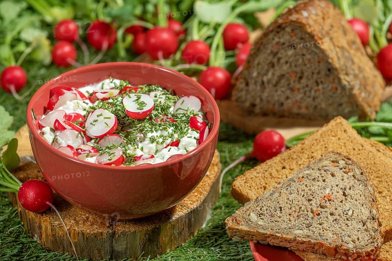 Bread next to a bowl of cream cheese, radishes and herbs on a green meadow