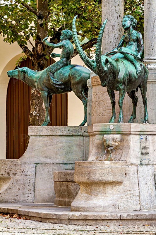 Bronze figures of riders on animals at an ornate fountain with arched structures, The fountain in the town of Kempten im Allgäu