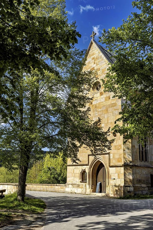 A Gothic church made of sandstone surrounded by trees under a bright blue sky, the Liborius Chapel on the Werra Bridge near Creuzburg in Thuringia