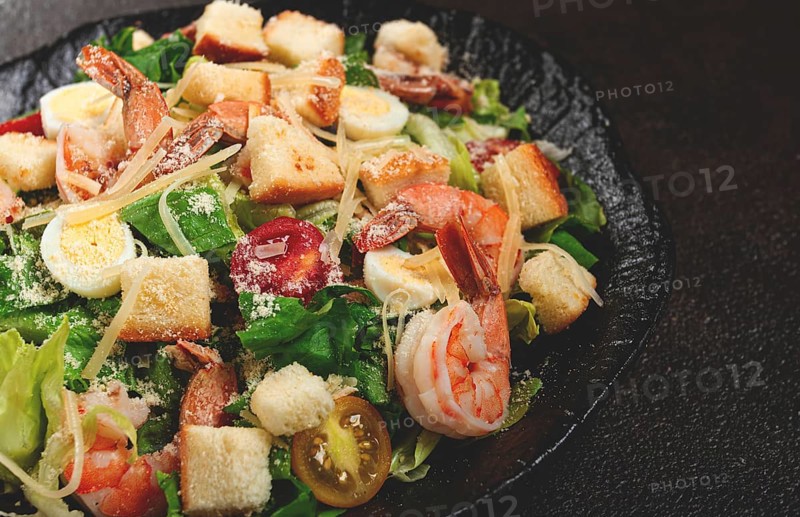 Caesar salad with tiger prawns, shrimp salad, lettuce leaves, quail eggs, cherry tomatoes, parmesan cheese, on a black plate