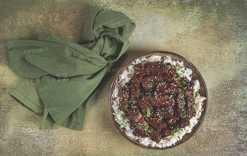 Crispy beef, with green onions and sesame seeds, on a rice pad, in a bowl with chopsticks, on a textured surface, no people