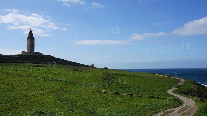 Coast with lighthouse and green field under a blue sky with clouds and a path to the sea, A Coruna, Spain