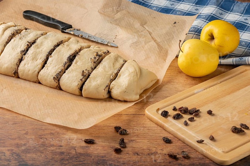 Dough pieces with poppy seed filling on baking paper, next to apples and raisins