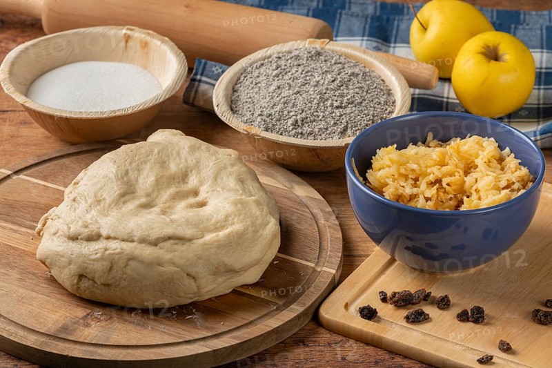 Dough prepared next to poppy seeds, grated apple and raisins on a wooden table