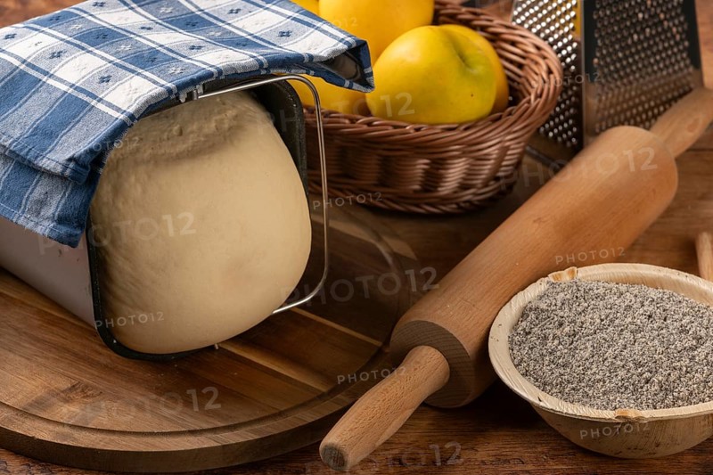 Dough in a metal mold under a tea towel next to apples, poppy seeds and rolling pin