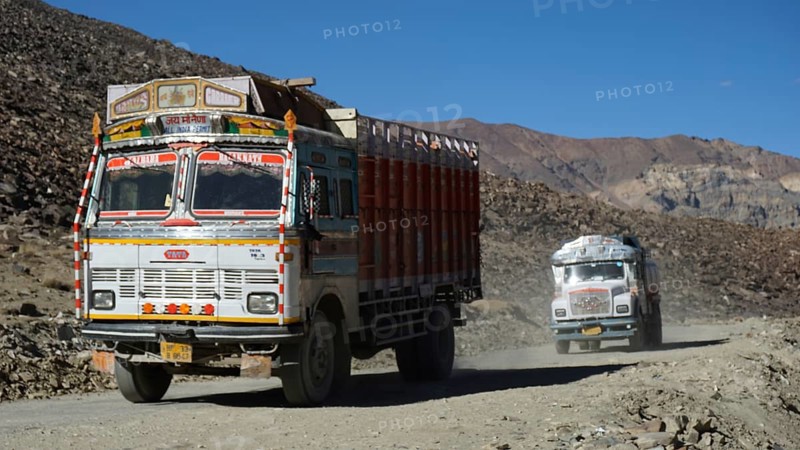 Two traditionally painted Indian trucks drive on a dusty, mountainous Manali-Leh Highway, Himachal Pradesh, Ladakh, India