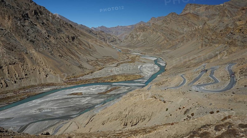 Shimmering blue Indus meanders through a barren valley with serpentines of a road leading down to the river under a blue sky, trekking in Ladakh, Himalayas, India