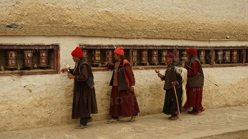 Four believers in traditional clothing turn a series of prayer wheels along the old wall of a Buddhist monastery, trekking in Ladakh, Himalayas, India