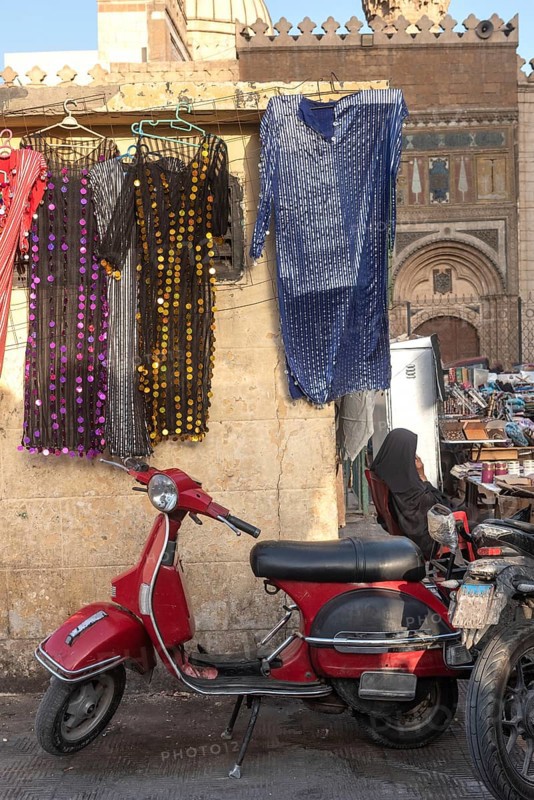 A cool looking red scooter parked amongst a busy street market selling colourful women's dresses near the famous Khan el Khalili Bazaar, historical Cairo, Egypt