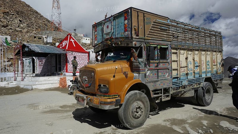 Colourful truck at Khardung-La Pass next to other vehicles and small houses with Indian flag under cloudy sky, trekking in Ladakh, Himalayas, India