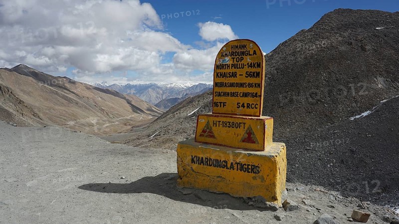 Signpost on dusty Khardung-la Pass in the mountains with sweeping views of surrounding mountains and cloudy skies, trekking in Ladakh, Himalayas, India