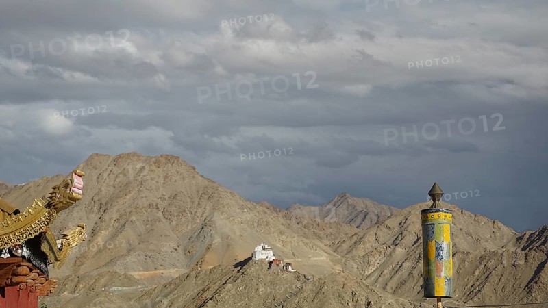 View towards Namgyal Tsemo monastery, dramatic sky with pediment of a Buddhist temple with Tibetan relief in the foreground of a prayer wheel against a mountain backdrop, Leh, trekking in Ladakh, Himalayas, India