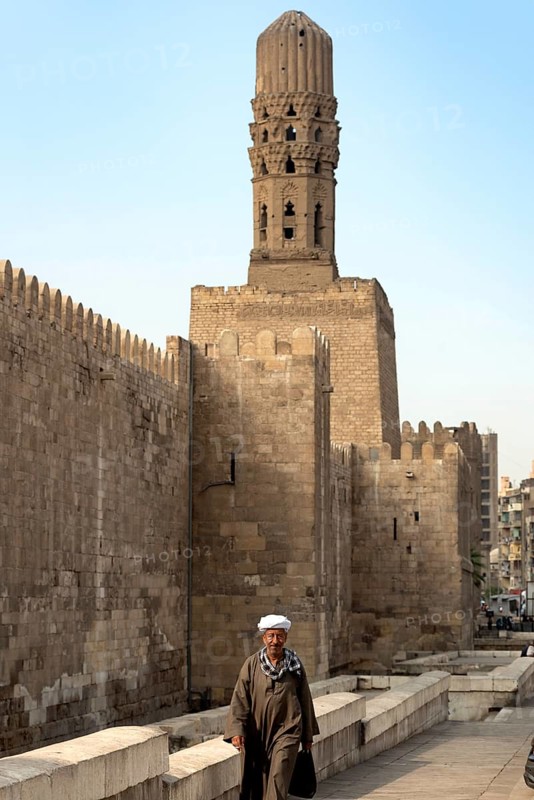 Cairo, Egypt, November 26th 2022 An Egyptian man wearing traditional clothes walking in front of the minaret of Al Hakim Mosque and the ancient city walls of Fatimid Islamic Cairo, Egypt