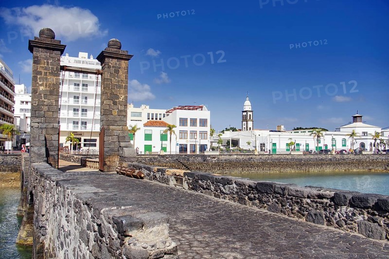 Historic stone drawbridge (Puente de las Bolas) over water with a view of a coastal town under a blue sky, Arrecife, Lanzarote, Canary Islands, Spain