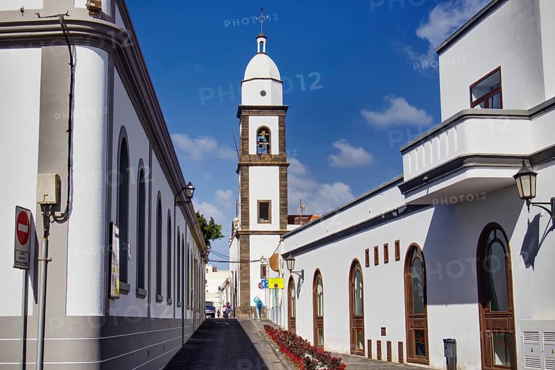 White buildings with church tower of the Iglesia de San Ginés church along a paved street under blue sky, Arrecife, Lanzarote, Canary Islands, Spain