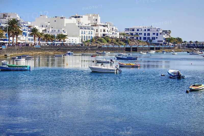 Blue water with boats in the Charco de San Ginés lagoon in front of a coastal town with palm trees and white buildings under clear skies, Arrecife, Lanzarote, Canary Islands, Spain