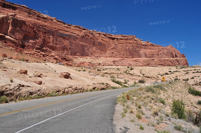 Road leading past a barren, red rock formation under bright blue sky, Arches National Park, Utah, USA