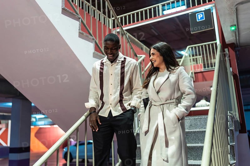 Diverse couple leaving an underground parking lot, laughing and enjoying their date night experience while descending concrete stairs with modern lighting and urban elements